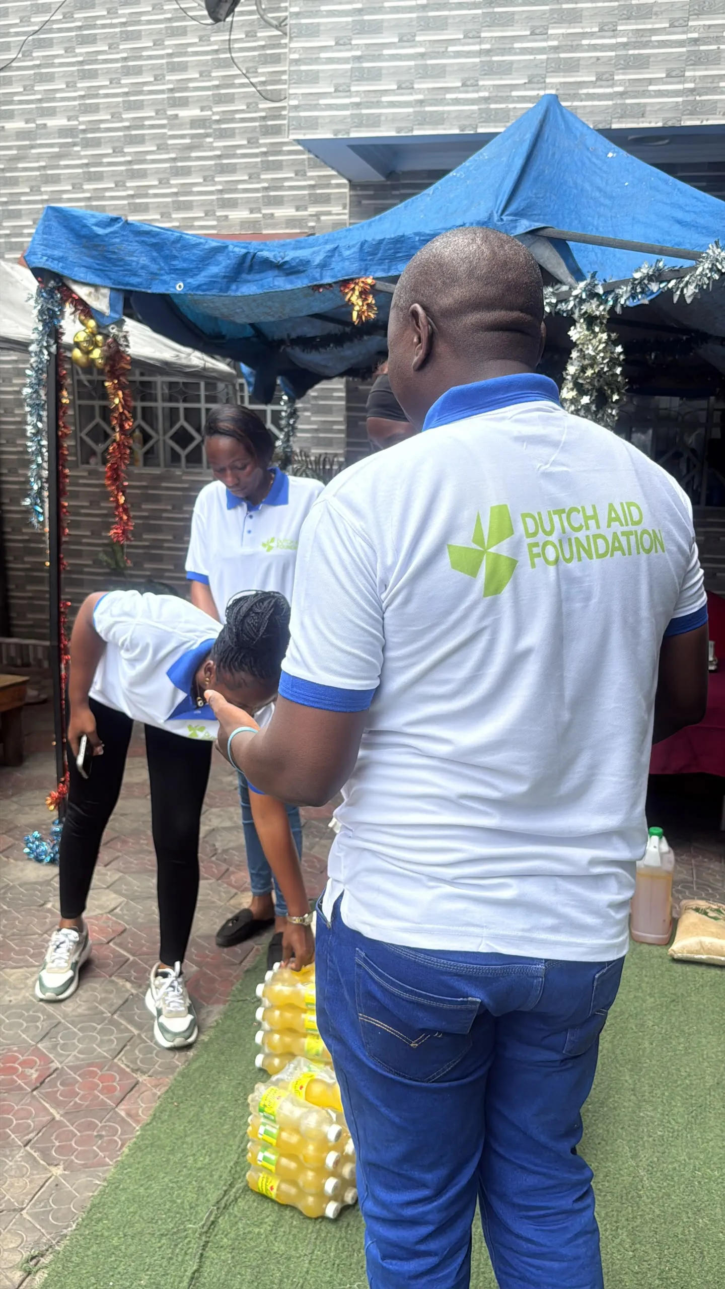 Een medewerker van Dutch Aid Foundation, van achteren gezien met het DAF-logo op het shirt, staat bij pakken flesjes drinken in Congo.