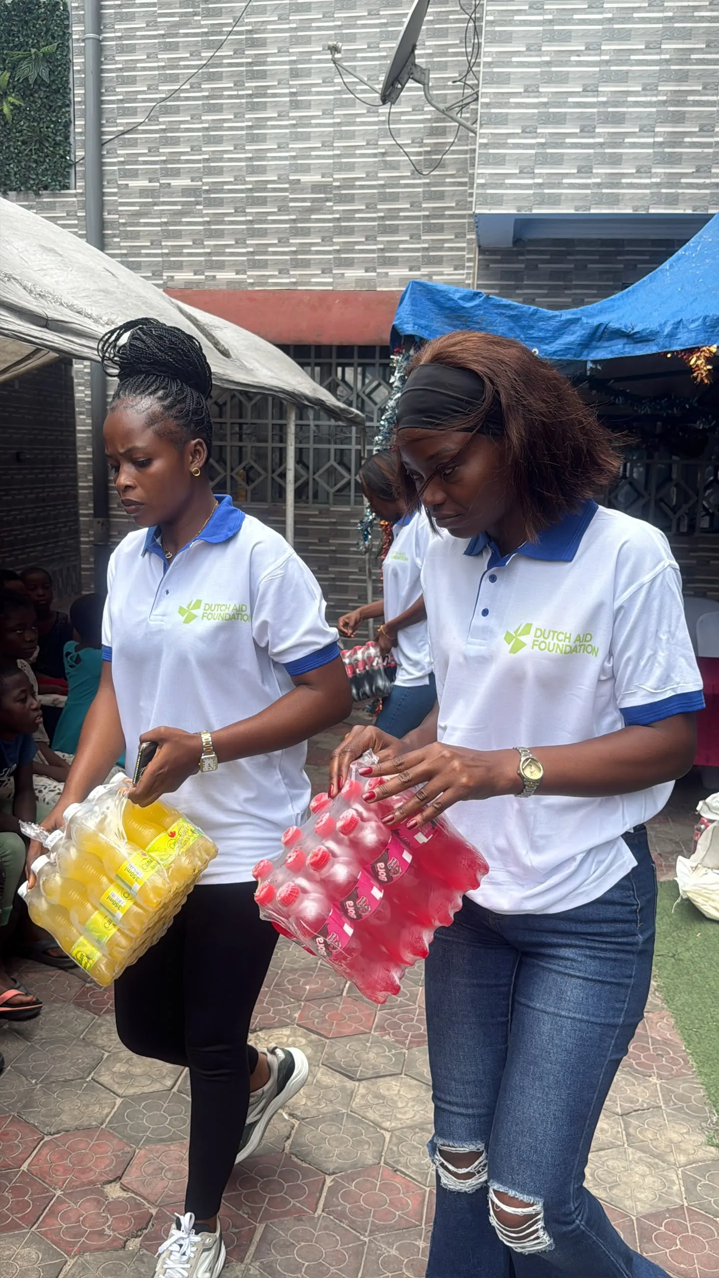 Twee medewerkers van Dutch Aid Foundation dragen pakken flesjes drinken over een binnenplaats in Congo.