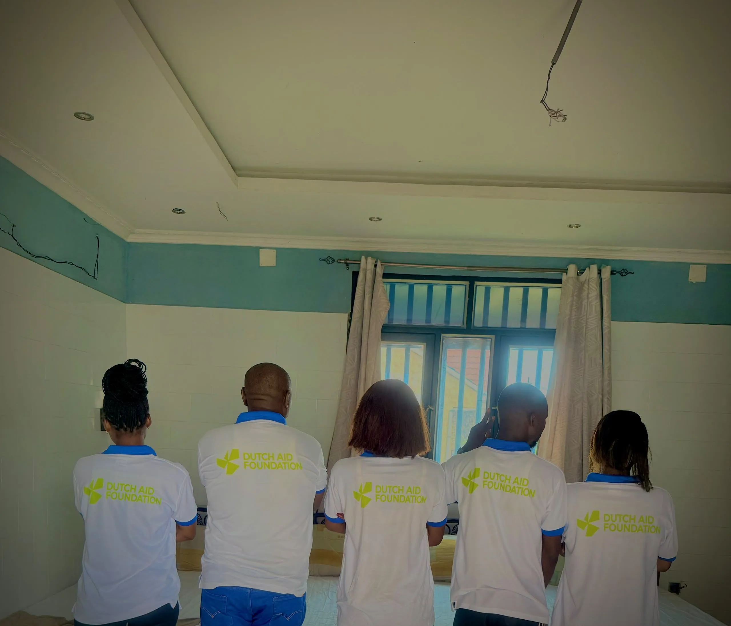Vijf medewerkers van Dutch Aid Foundation, van achteren gezien in hun DAF-shirts, in een ruimte in Congo.
