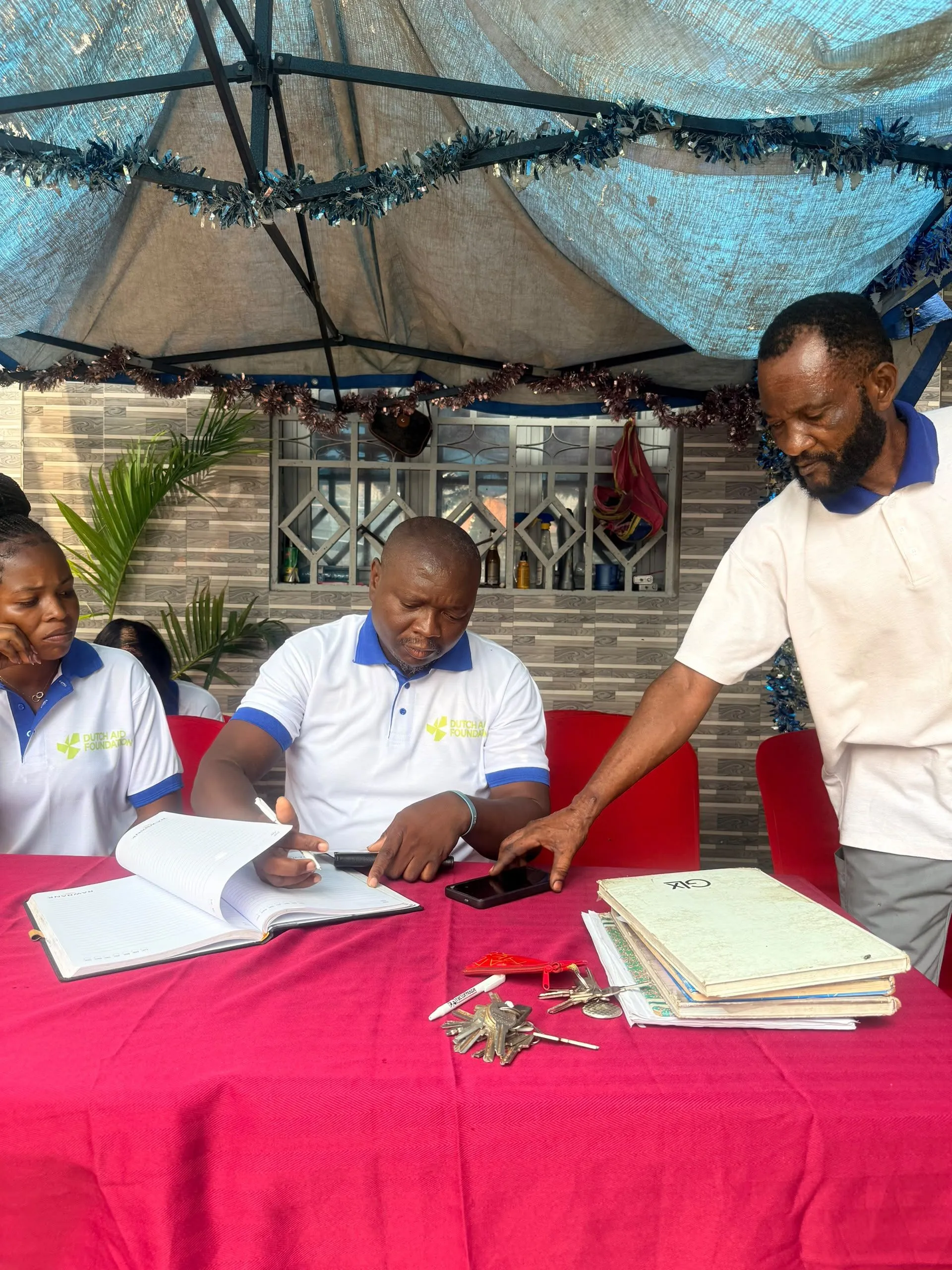 Medewerkers van Dutch Aid Foundation aan een tafel met een rood kleed; één schrijft in een notitieboek terwijl een ander toekijkt, in Congo.